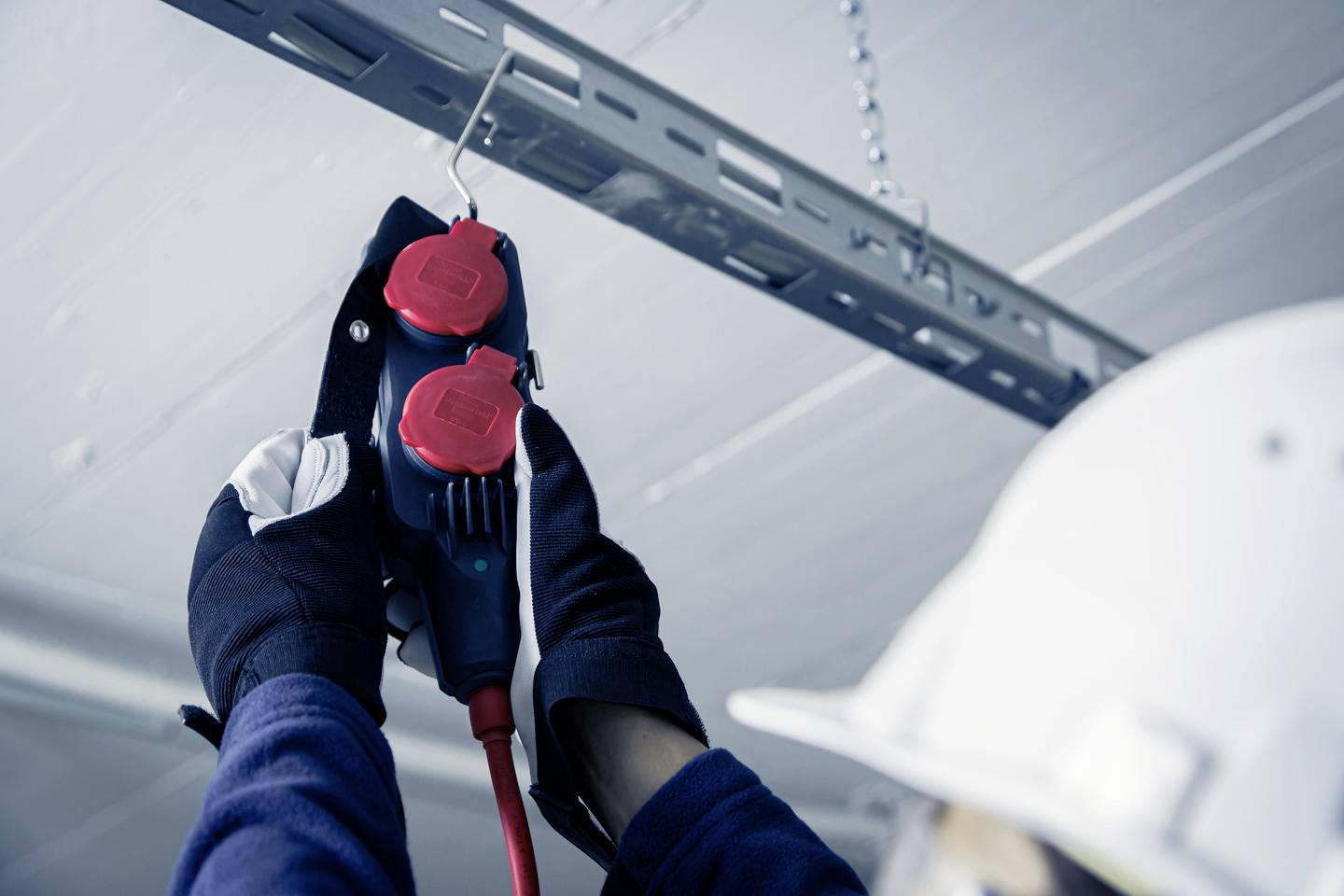 Eine Person mit Schutzhelm und Handschuhen installiert eine rote Steckdose an einer Deckenhalterung.