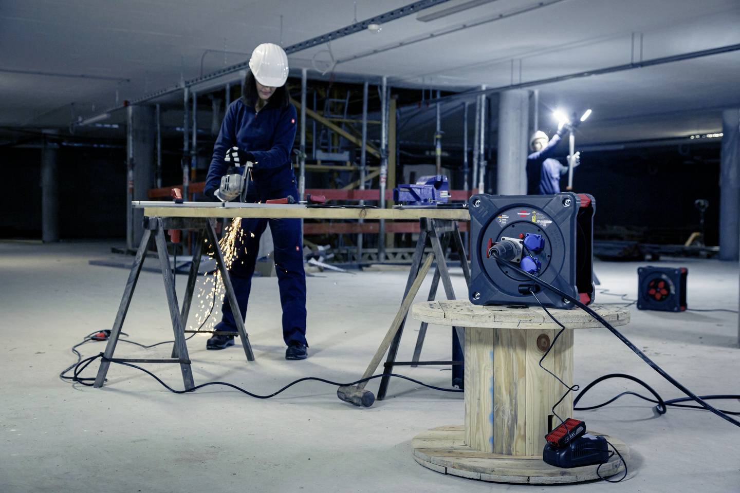 Eine Person schneidet Metall mit einem Winkelschleifer auf einer Baustelle. Funken fliegen, ein zweiter Arbeiter beleuchtet die Szene.