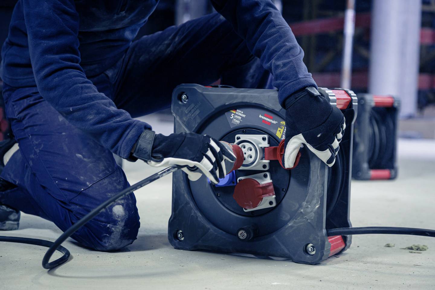 Eine Person mit Handschuhen schließt ein Stromkabel an eine Kabeltrommel auf einer Baustelle an.