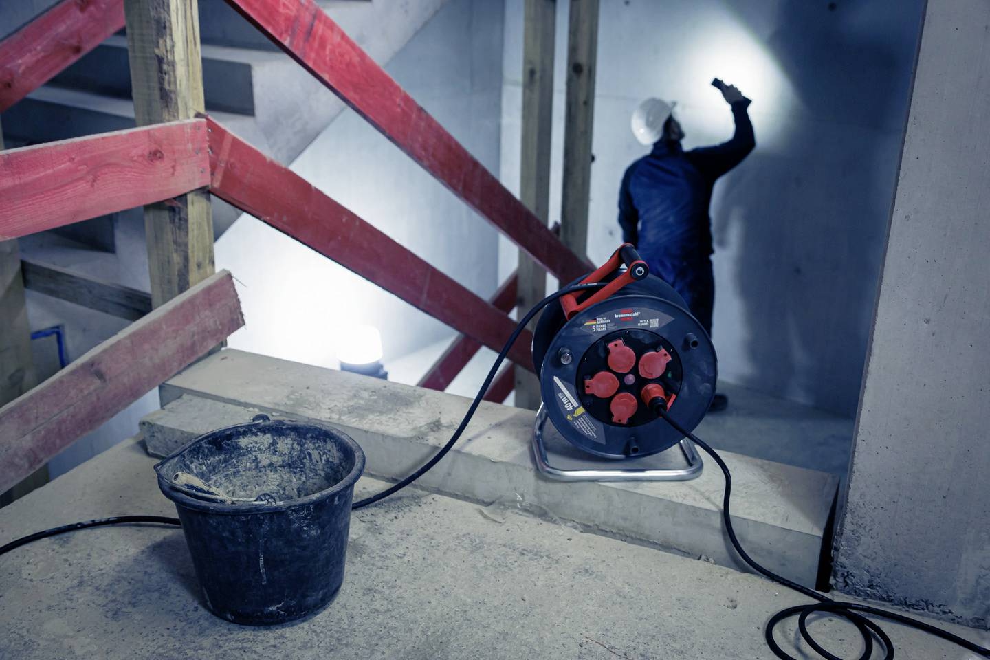 Ein Arbeiter in blauer Schutzkleidung und Helm untersucht eine Baustelle mit einer Taschenlampe. Im Vordergrund steht ein Eimer und ein Kabeltrommel.
