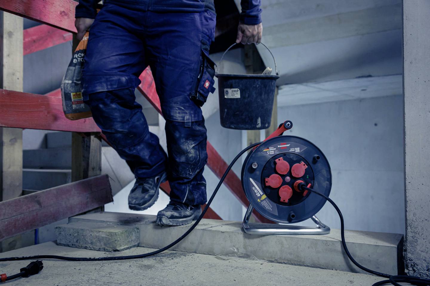 Eine Person trägt zwei Eimer auf einer Baustelle. Im Vordergrund befindet sich eine Kabeltrommel. Es scheint sich um ein Arbeitsumfeld zu handeln.