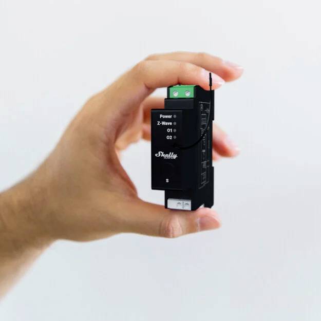 Eine Hand hält ein schwarzes, rechteckiges elektronisches Gerät. Auf der Vorderseite des Geräts stehen die Wörter 'Power' und 'Shelly'.