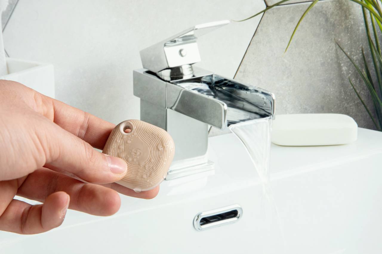 Hand hält ein kleines, quadratisches Objekt vor einem Wasserhahn, aus dem Wasser fließt. Im Hintergrund sind Pflanzen zu sehen.