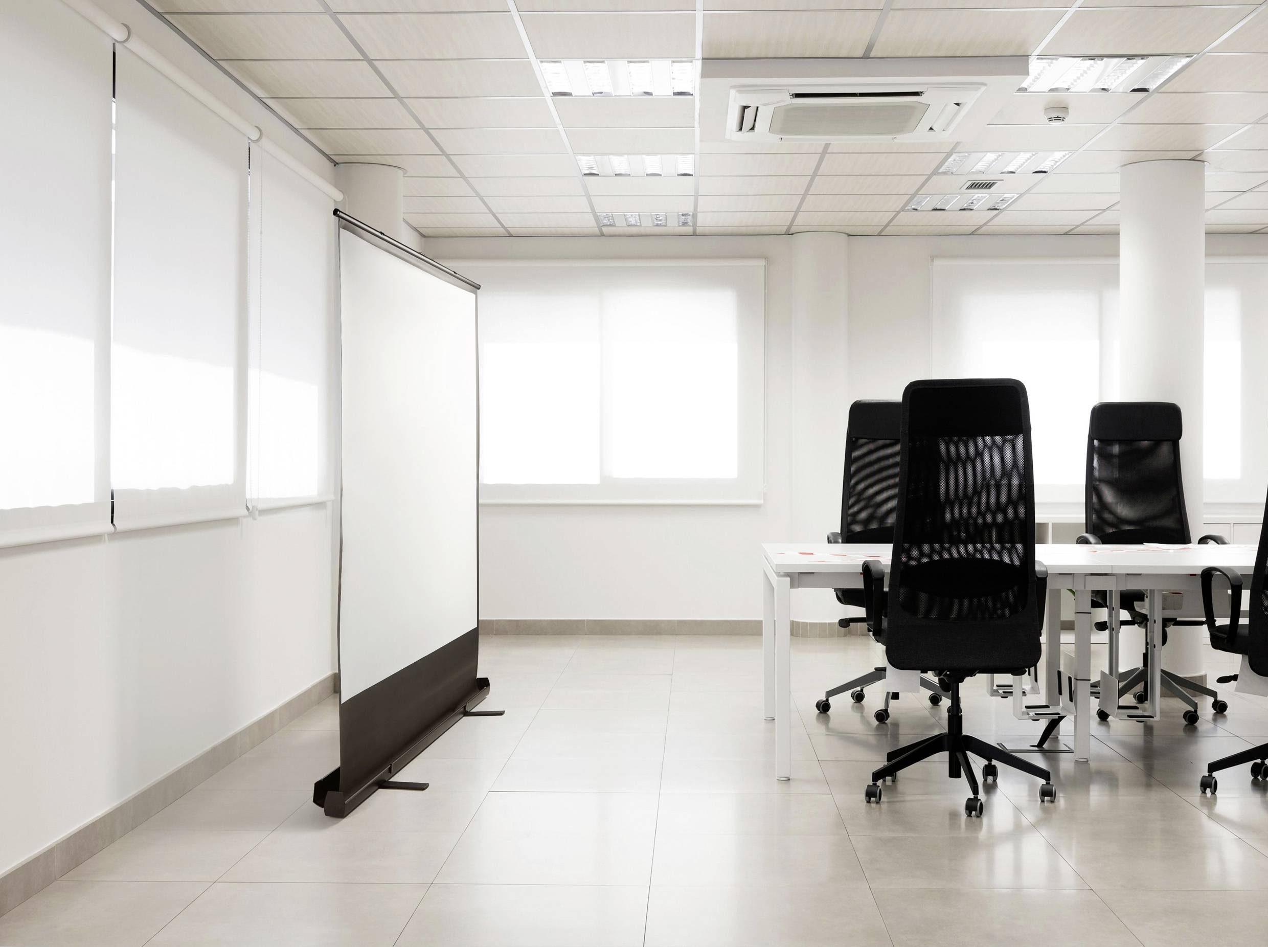 Ein heller Büroraum mit einem leeren Projektorbildschirm links und drei schwarzen Bürostühlen an weißen Tischen rechts.