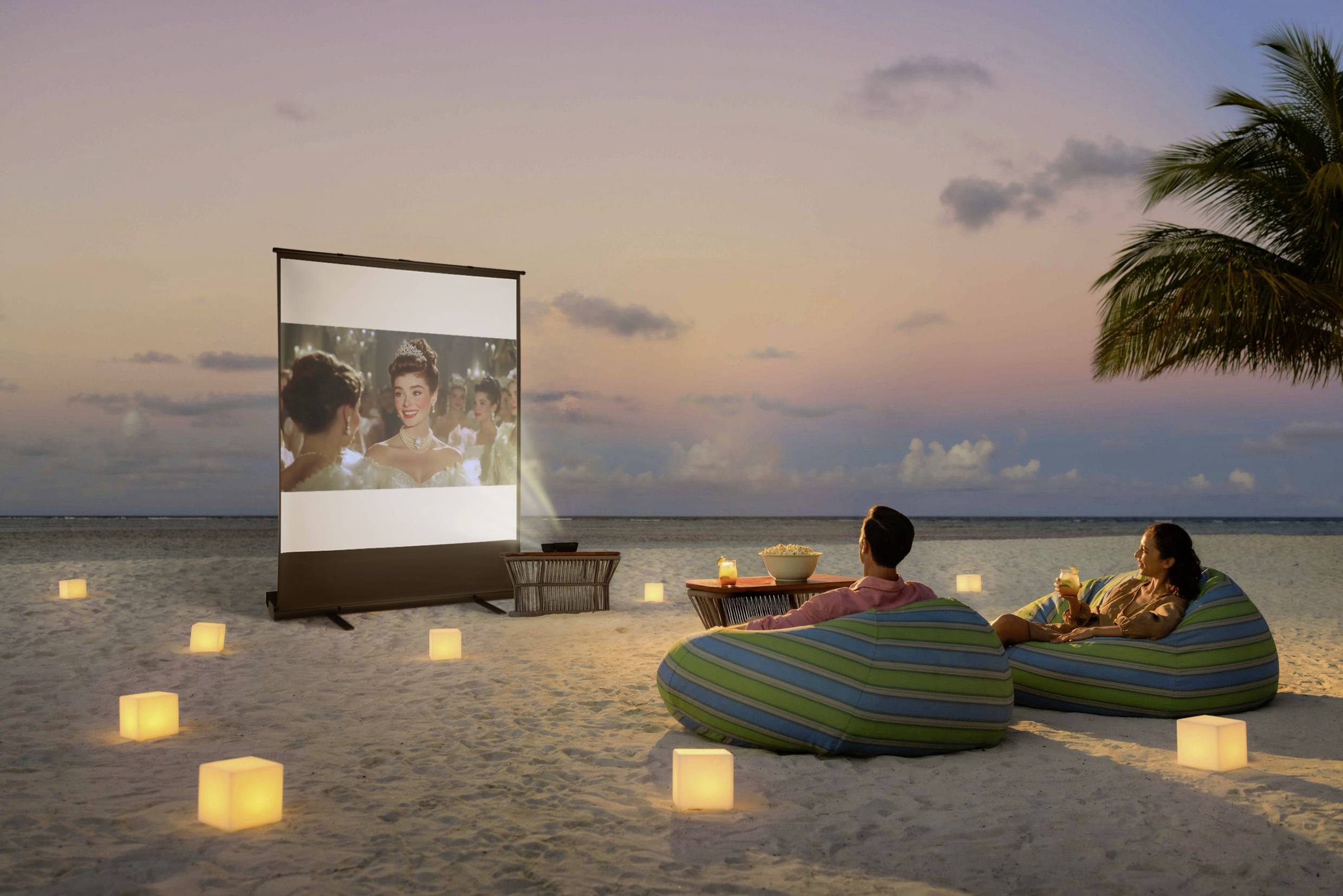Ein Paar sitzt auf Sitzsäcken am Strand und schaut einen Film auf einer Leinwand bei Sonnenuntergang, umgeben von Laternen.