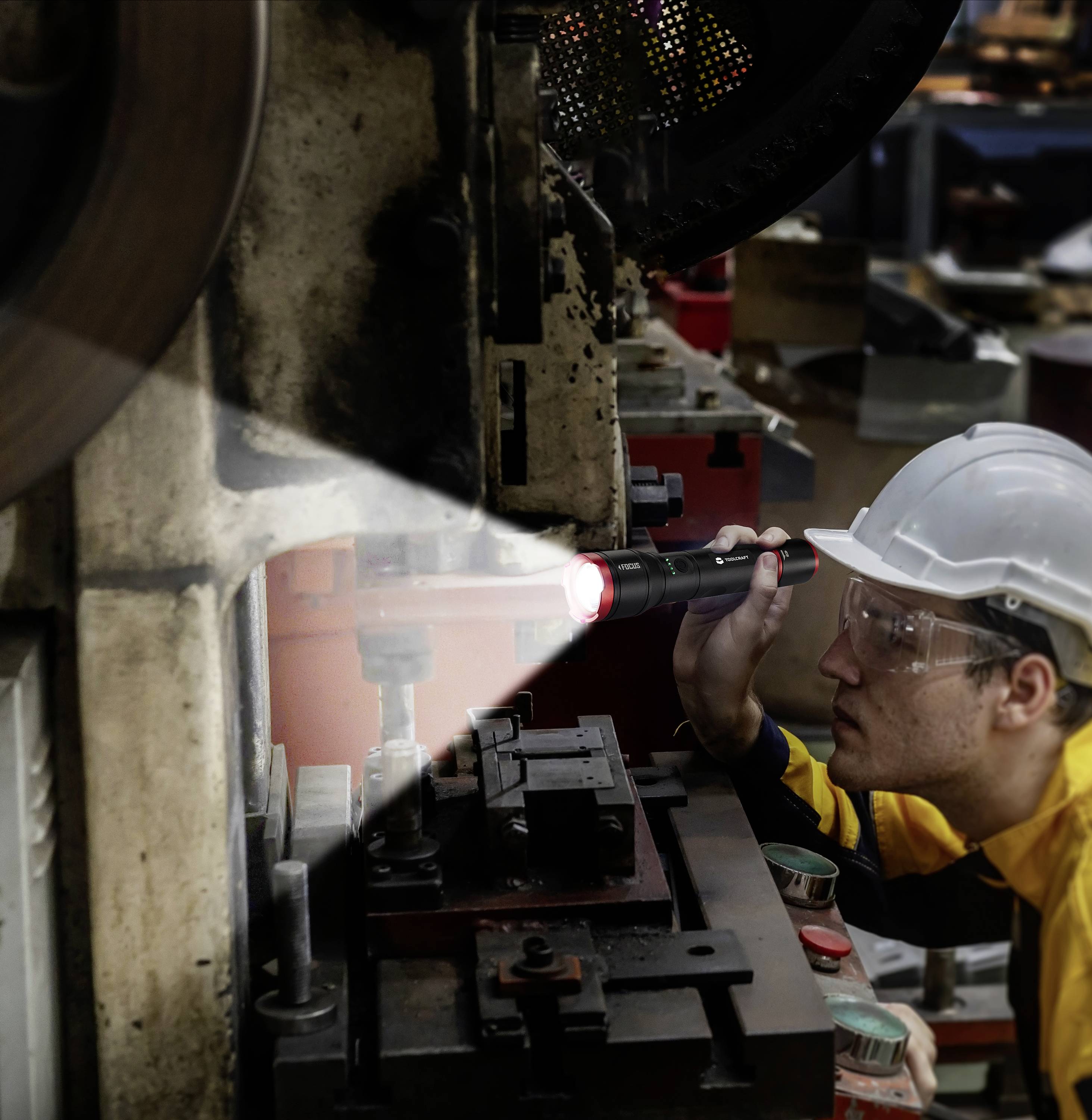 Ein Arbeiter mit Schutzhelm und Brille inspiziert eine industrielle Maschine mit einer Taschenlampe in einer Fabrikumgebung.