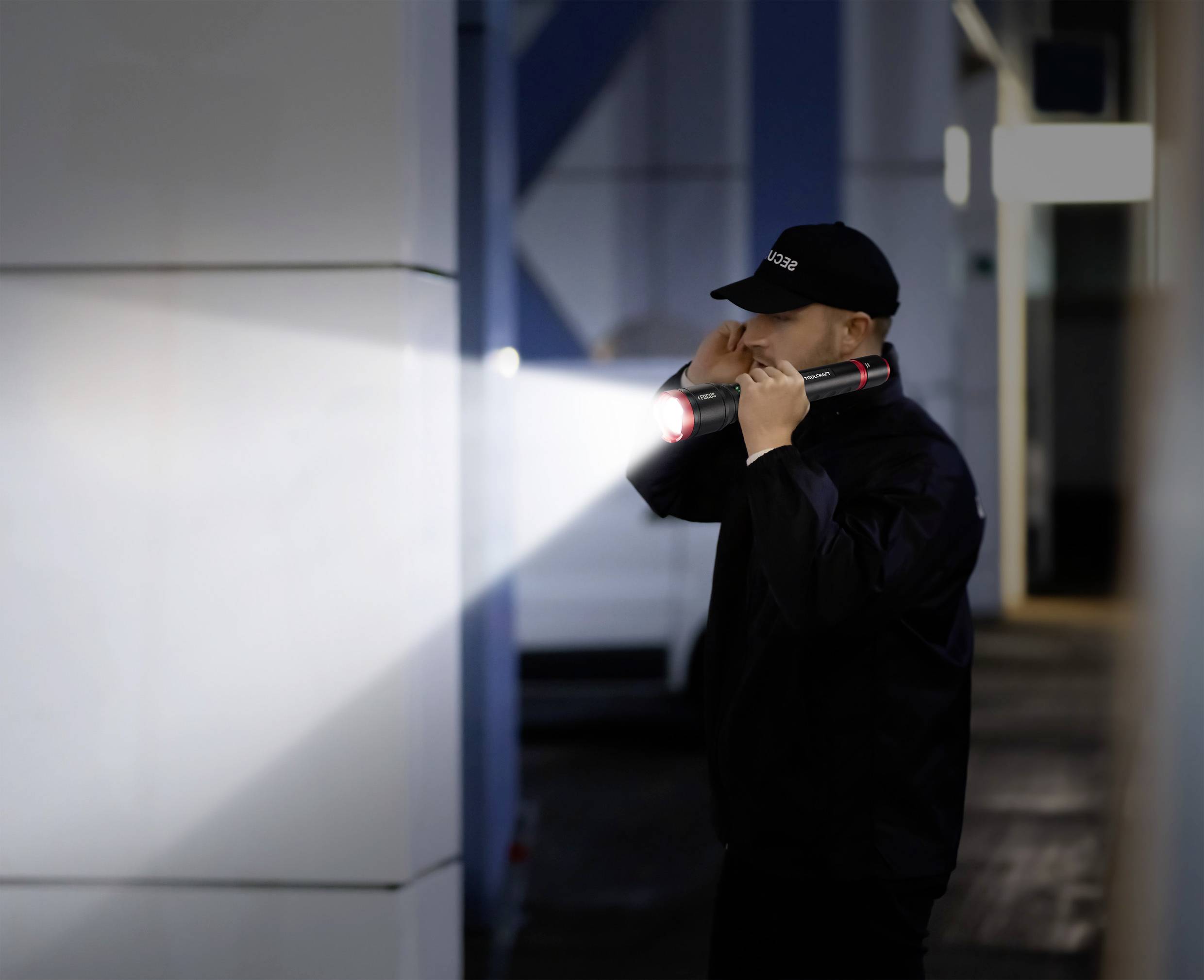 Ein Sicherheitsmitarbeiter inspiziert mit einer Taschenlampe einen dunklen Bereich. Trägt Uniform und Mütze, Umgebung ist industrieähnlich.