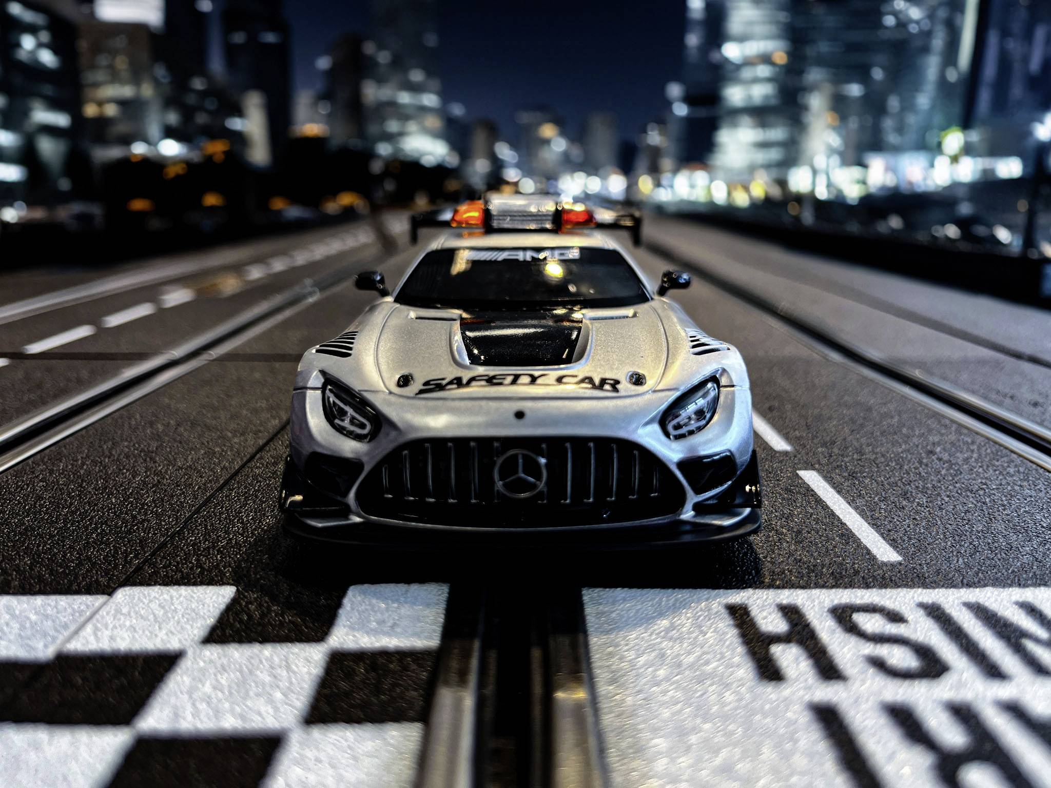 Ein Modellautorennauto mit der Aufschrift 'Safety Car' fährt auf einer beleuchteten Rennstrecke vor einer verschwommenen Stadtlandschaft.