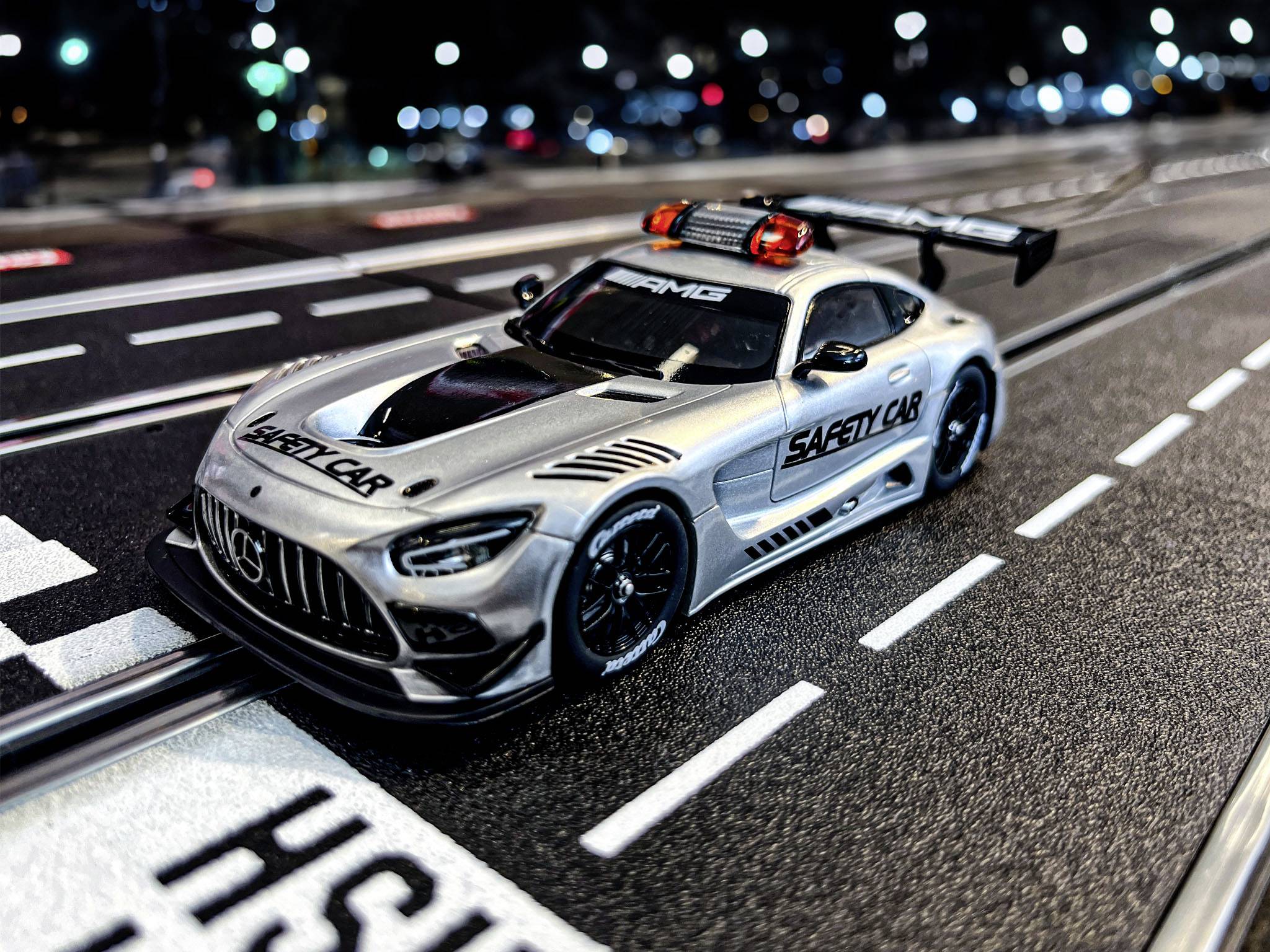 Ein silbernes Spielzeugauto mit der Aufschrift 'Safety Car' steht auf einer Rennstrecke bei Nacht. Beleuchtete Gebäude im Hintergrund.