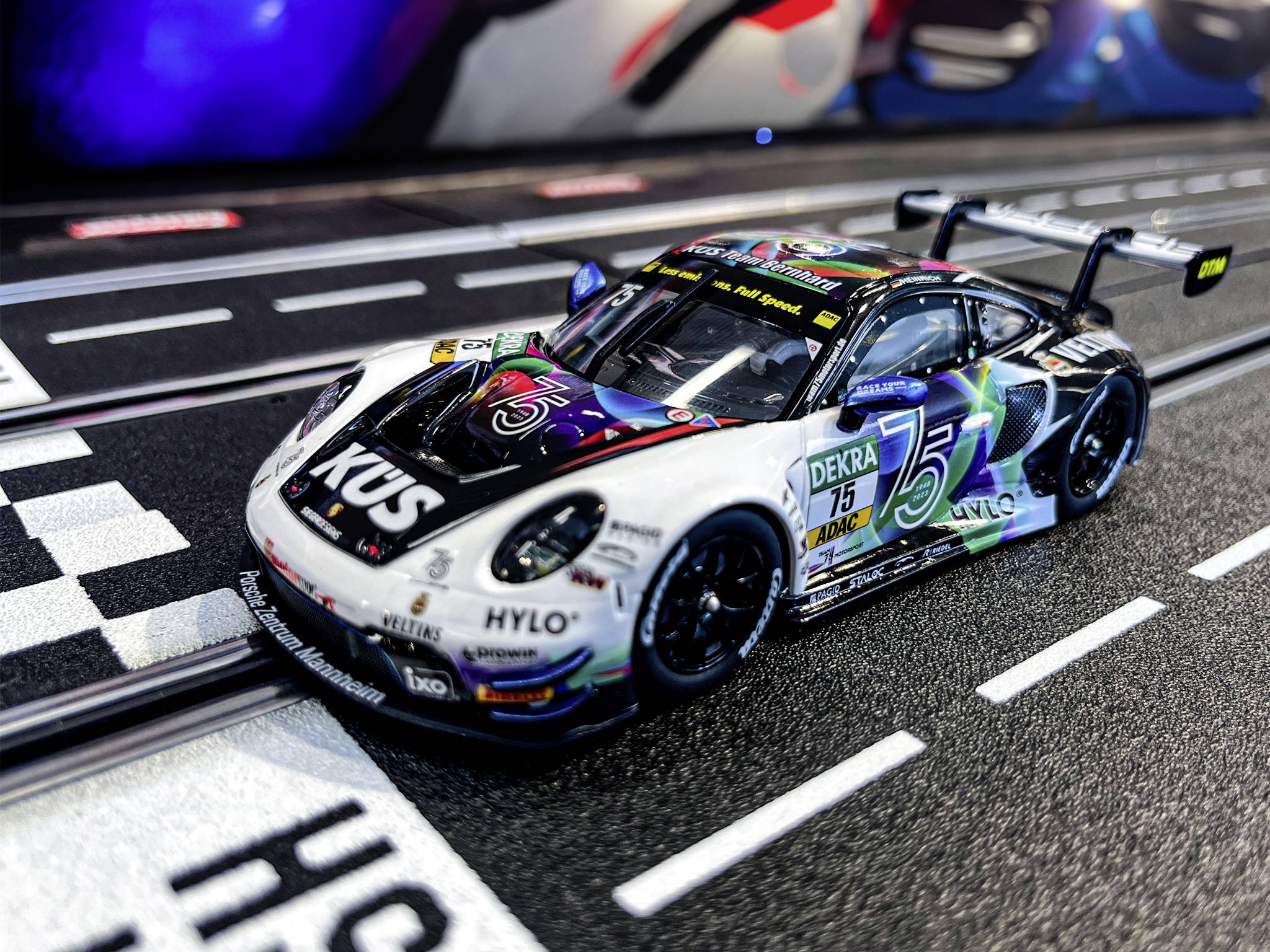 Ein Spielzeugauto in Rennlackierung steht auf einer Slotcar-Rennbahn, bereit zum Start. Auf der Motorhaube steht 'KÜS'.