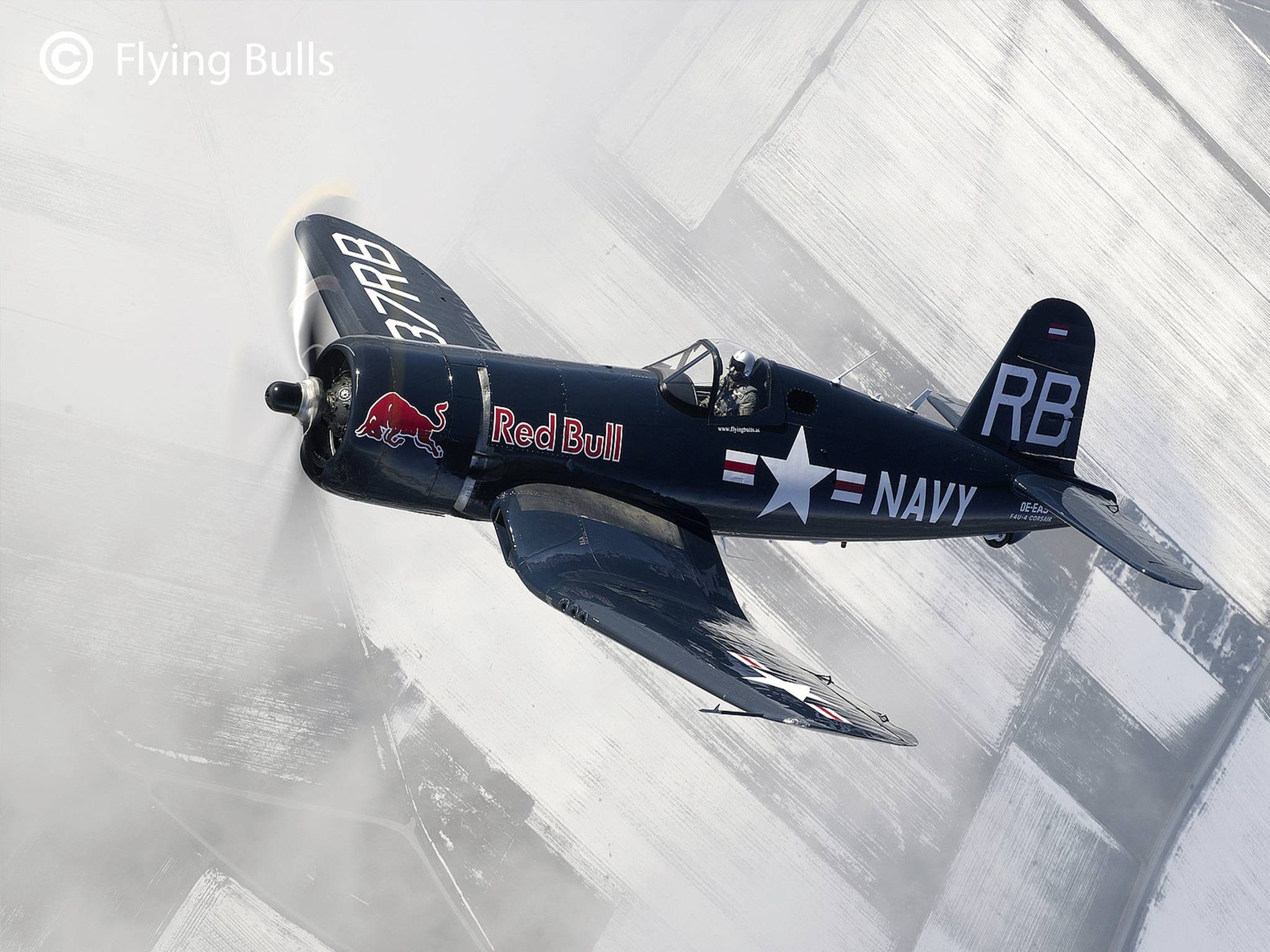 Ein dunkles Kampfflugzeug mit der Aufschrift 'Red Bull' und 'NAVY' fliegt in einer Rauchwolke über einer verschneiten Landschaft.