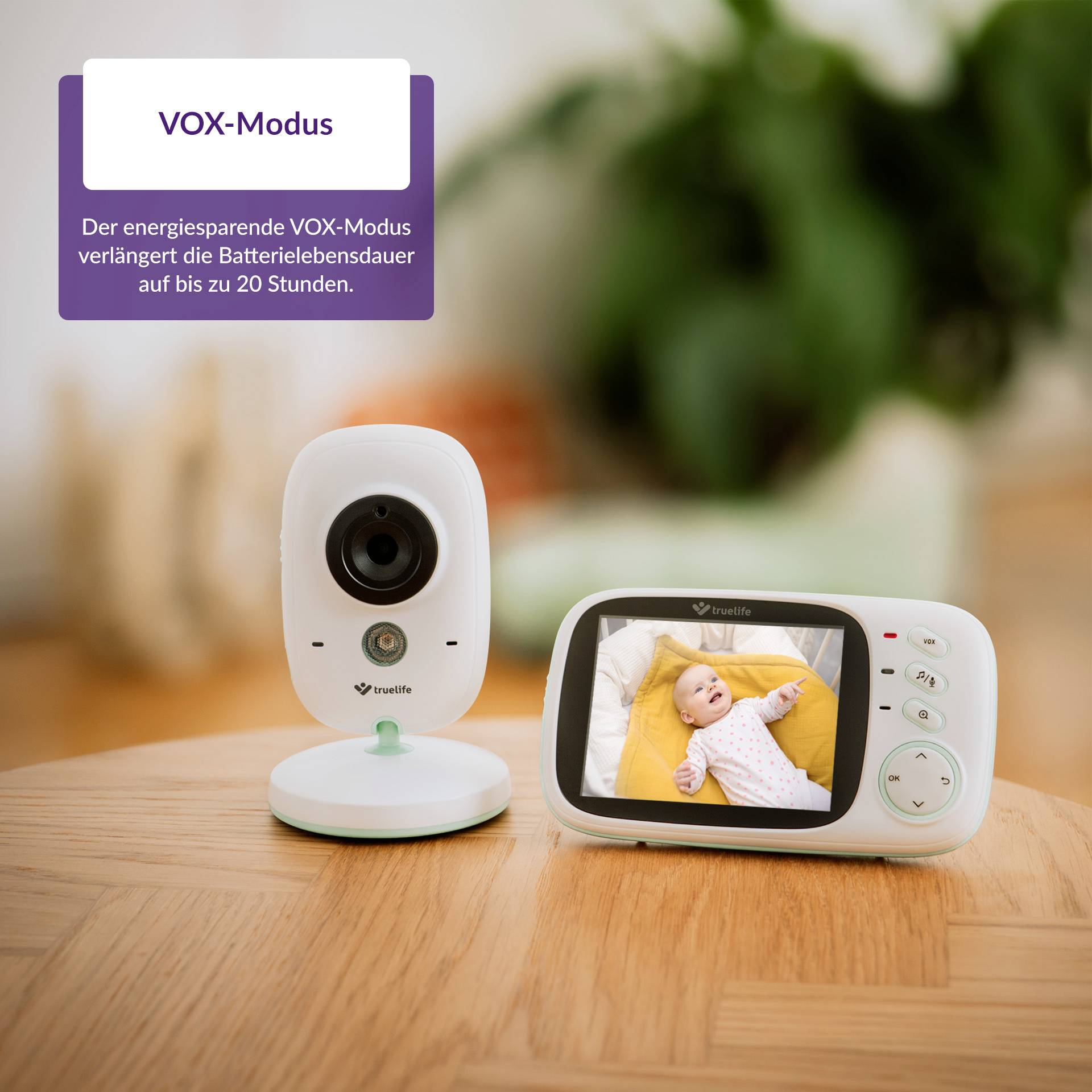Babyphone auf Holztisch, Kamera links, Monitor rechts zeigt Baby. Text betont energiesparenden VOX-Modus mit bis zu 20 Stunden Akkulaufzeit.