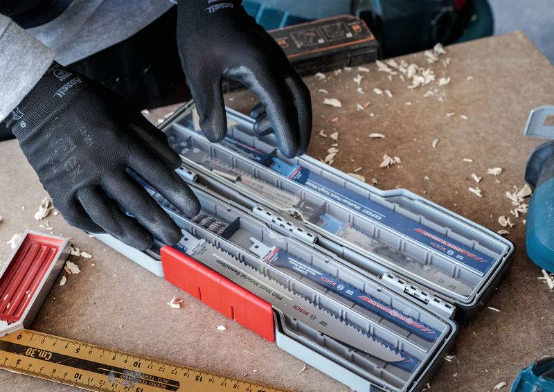 Eine Person mit schwarzen Handschuhen öffnet einen Kasten mit verschiedenen Sägeblättern auf einer Werkbank, umgeben von Holzspänen.
