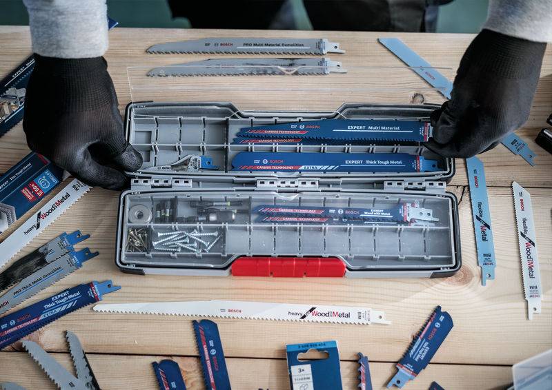 Eine Person sortiert Sägeblätter in einem offenen Werkzeugkasten auf einem Holztisch. Verschiedene Sägeblätter sind herum verteilt.