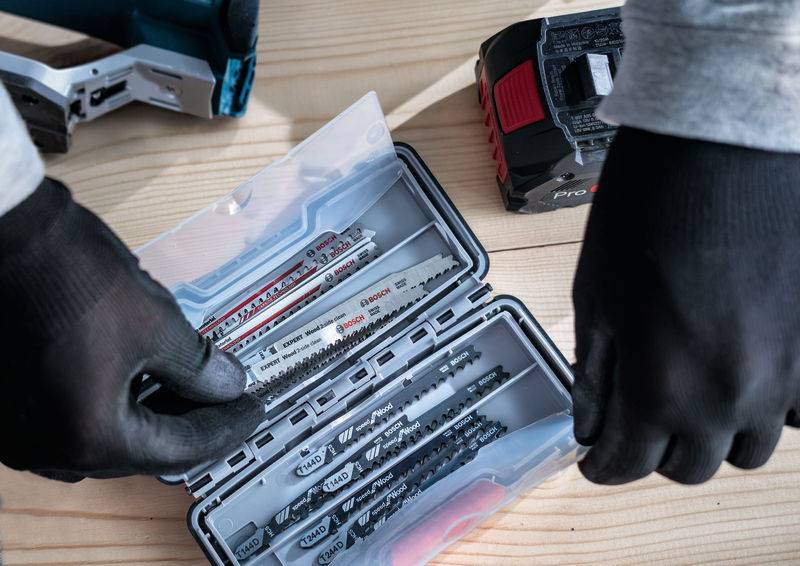 Eine Person in Handschuhen wählt eine Sägeklinge aus einer Kunststoffbox auf einem Holztisch, mit einer Akku-Sägemaschine im Hintergrund.