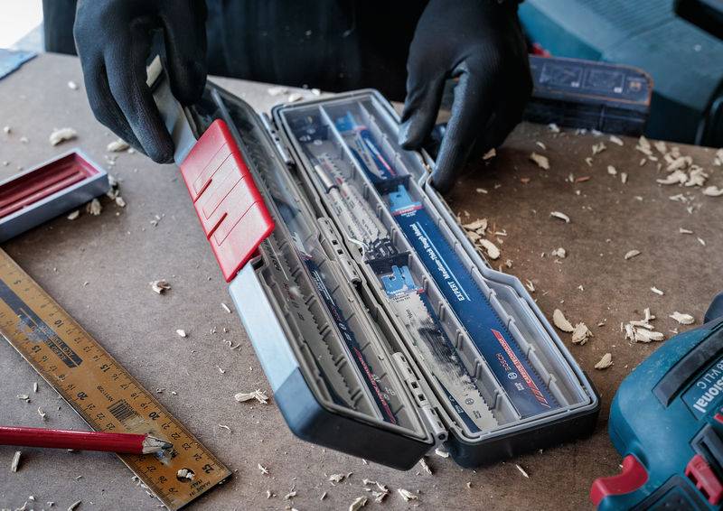 Eine Person mit schwarzen Handschuhen öffnet eine Bohrerkassette auf einer Werkbank. Umgeben von Holzspänen, Lineal und Werkzeugen.