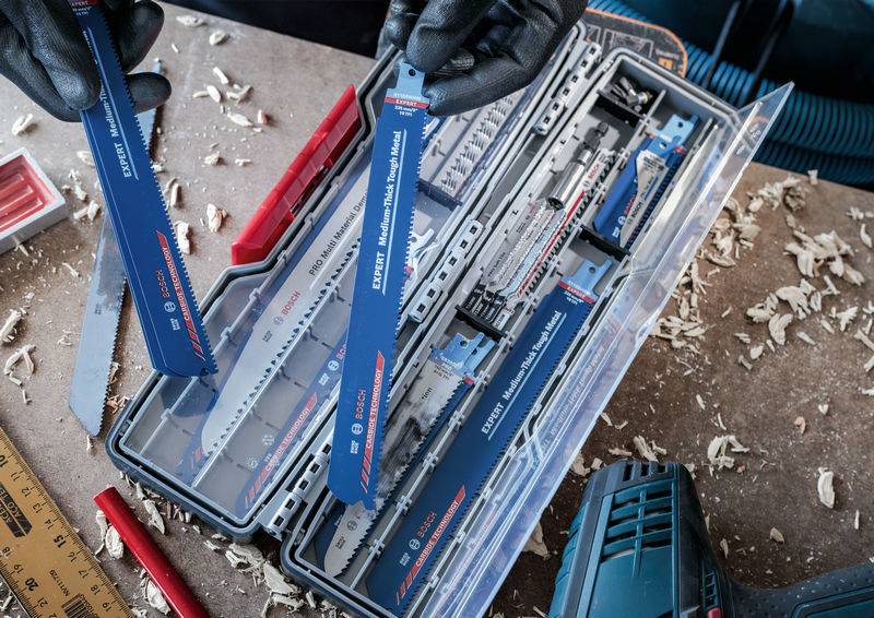 Eine Person in schwarzen Handschuhen hält ein blaues Säbelsägeblatt neben einer offenen Kunststoffbox mit mehreren Sägeblättern auf einer Werkbank.