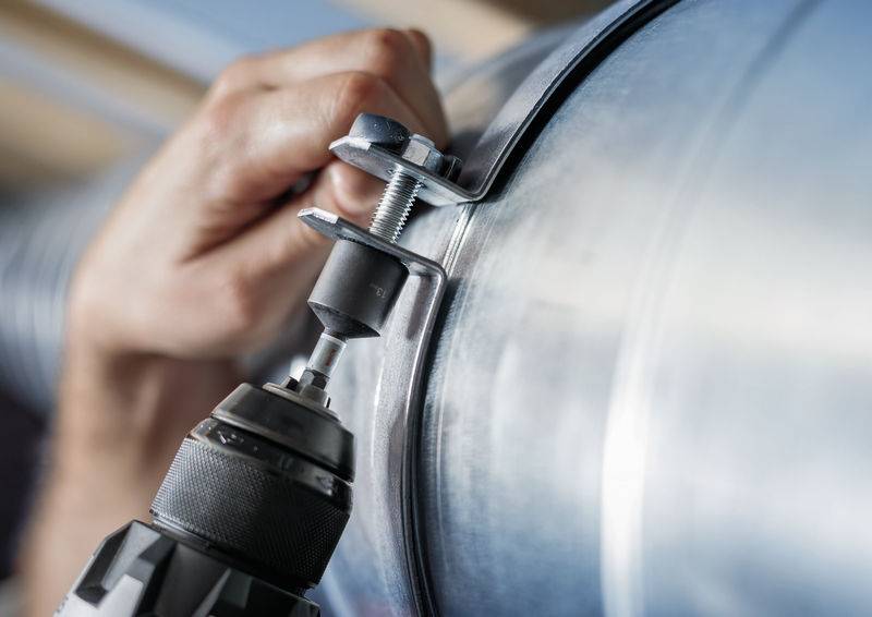 Eine Nahaufnahme einer Hand, die mit einer Bohrmaschine arbeitet, um eine Metallklemme an einem großen Metallrohr zu befestigen.