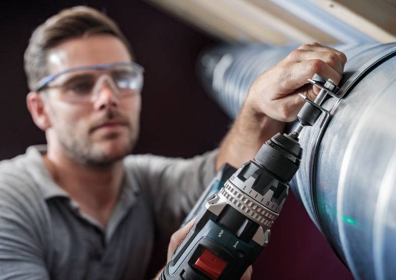Eine Person mit Schutzbrille montiert ein großes Metallrohr mit einem Bohrschrauber in einem industriellen Umfeld.