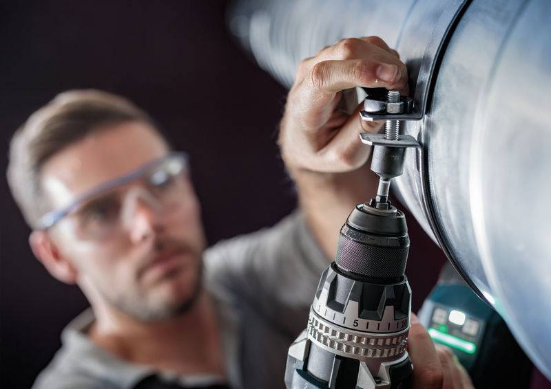 Ein Mann trägt Schutzbrille und benutzt einen Bohrer, um an einem großen Metallrohr zu arbeiten. Fokus liegt auf dem Werkzeug und der Hand.