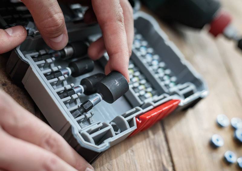 Person legt ein Werkzeugteil in eine Box mit mehreren Aufsätzen auf einem Holztisch. Fokus liegt auf der Ordnung der Werkzeuge.