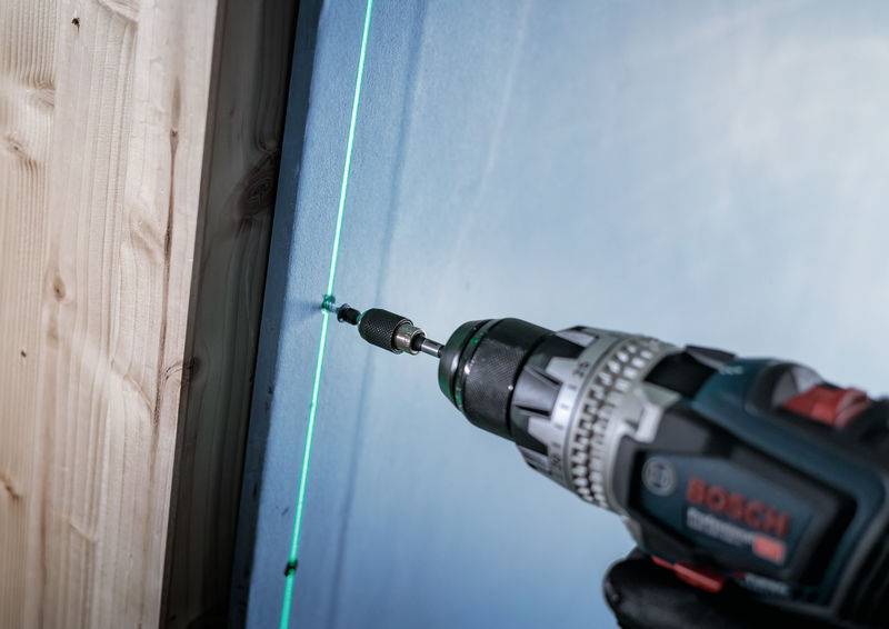 Eine Bohrmaschine mit einem grünen Laser richtet Schrauben auf einer Wand für präzise Bohrungen aus. Eine Holzplatte ist links sichtbar.