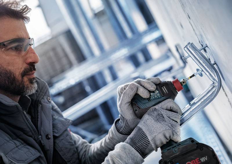 Eine Person bohrt mit einem Akkuschrauber ein Rohr an eine Wand. Sie trägt Schutzbrille und Handschuhe, arbeitet konzentriert.