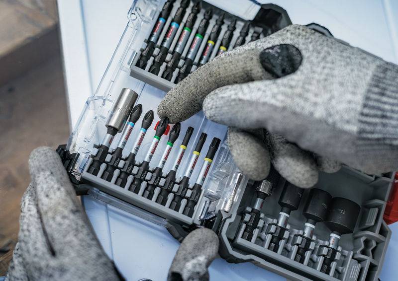 Eine Person mit grauen Handschuhen wählt einen Schraubendreher-Aufsatz aus einer offenen Box mit verschiedenen Bits.