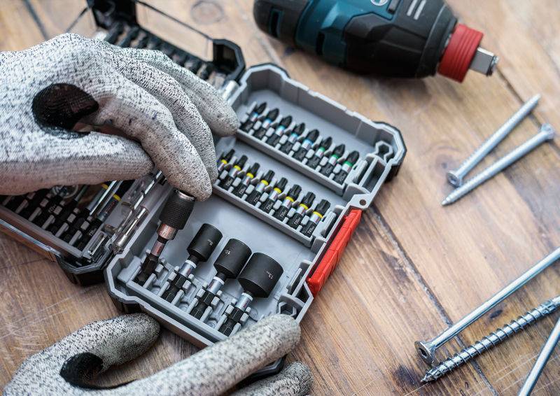 Eine Person mit Arbeitshandschuhen sortiert Schraubendreher-Bits in einem Kasten. Eine Bohrmaschine und Schrauben liegen daneben.