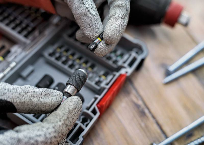 Zwei Hände in Arbeitshandschuhen halten einen Bohrer und einen Bohrkopf vor einer offenen Werkzeugkiste auf einem Holztisch.