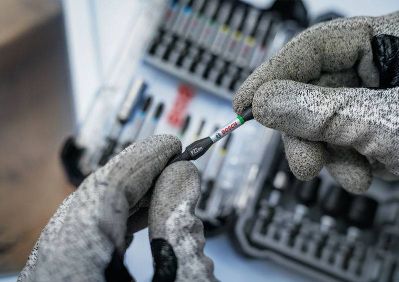 Eine Person mit grauen Handschuhen hält einen Bosch-Schraubendreher-Bit vor einem unscharfen Werkzeugset.