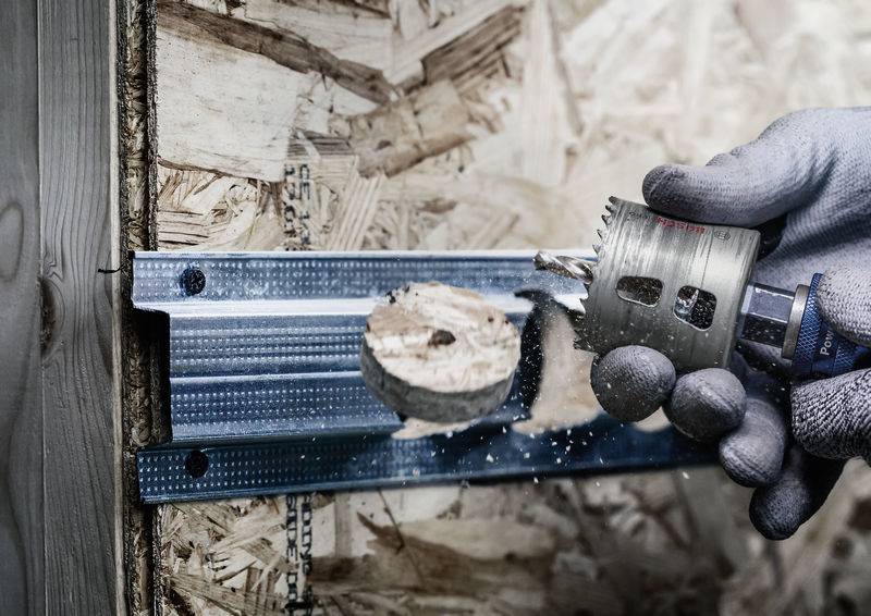 Eine Hand hält einen Bohrer, der ein rundes Loch in eine OSB-Platte bohrt, die an einem Metallprofil befestigt ist.