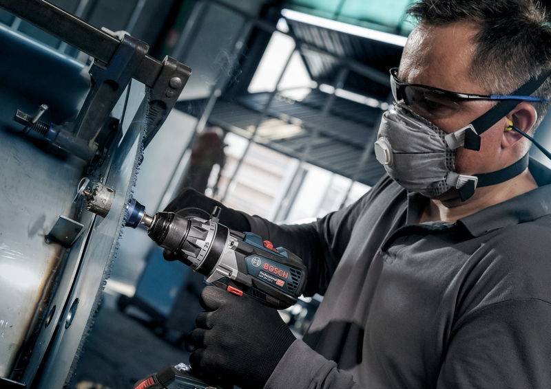 Ein Arbeiter mit Schutzbrille und Atemmaske benutzt eine elektrische Bohrmaschine an einem Metallstück in einer industriellen Umgebung.