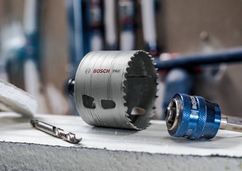 Bohreraufsätze und Zubehör liegen auf einer Werkbank. Im Fokus ist ein silberner, zylinderförmiger Sägeaufsatz mit 'BOSCH PRO'-Aufschrift.