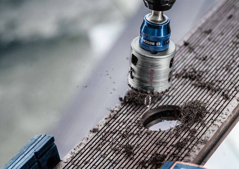 Ein Bohrer durchdringt mit einer Lochsäge ein Holzbrett, Späne sind um das Bohrloch herum verteilt.