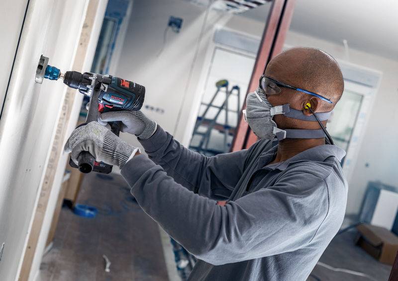 Person mit Schutzbrille, Ohrstöpseln und Atemmaske verwendet eine Bohrmaschine an einer Wand in einem Bau- oder Renovierungsbereich.