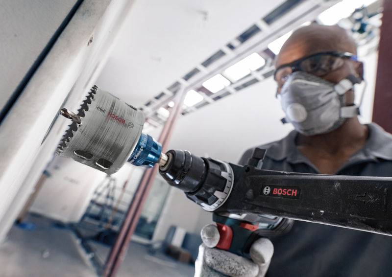 Eine Person trägt Schutzbrille und Maske, benutzt einen Bosch-Bohrer mit Lochsägeaufsatz, um ein Loch in eine Wand zu bohren. Hintergrund zeigt Baustelle.