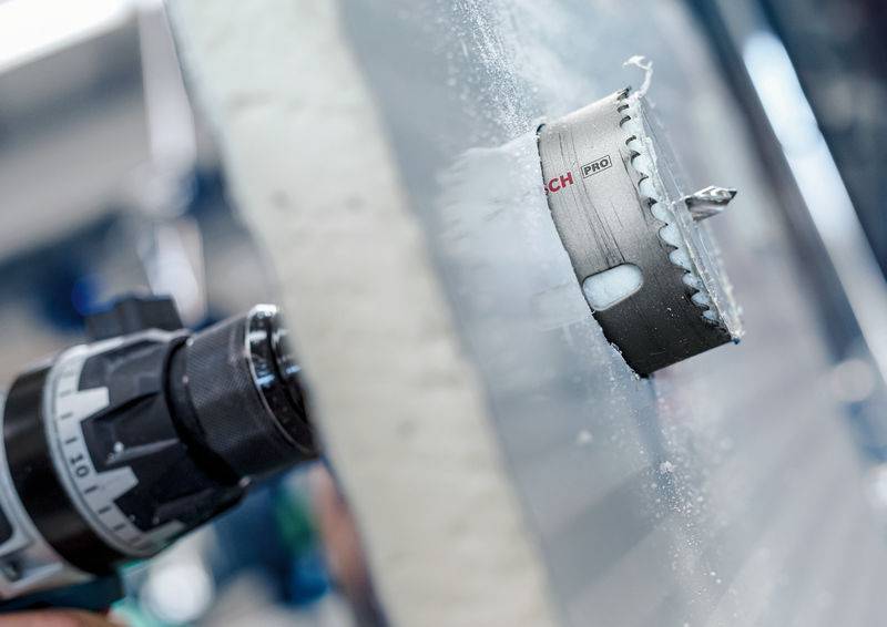 Eine elektrische Bohrmaschine mit einer Lochsäge bohrt in eine Mauer, Metallspäne fallen ab. Die Szene zeigt handwerkliche Arbeit.