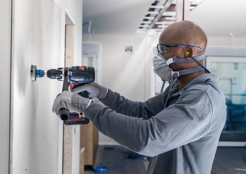 Ein Arbeiter mit Schutzbrille und Maske bohrt ein Loch in eine Wand auf einer Baustelle, mit einem blauen Akkubohrer in der Hand.