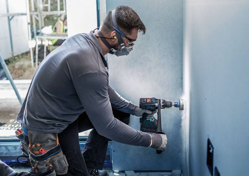 Ein Handwerker kniet in einem Raum und bohrt ein Loch in eine Wand mit einem Elektrowerkzeug. Er trägt Schutzbrille und Mundschutz.
