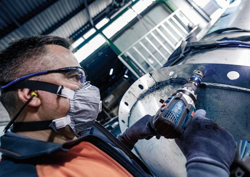 Ein Arbeiter trägt Schutzbrille und Maske, während er mit einem Elektrowerkzeug an einem großen Metallrohr in einer Fabrik arbeitet.