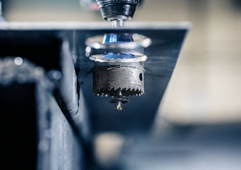 Eine Metallbohrmaschine bohrt ein rundes Loch in eine Metallplatte. Der Bohrkopf und entstehende Späne sind sichtbar.