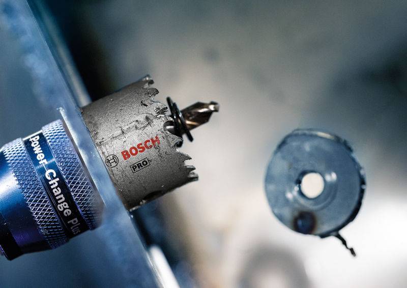 Bohrmaschine mit Lochsäge-Aufsatz der Marke Bosch PRO, bohrt ein Loch in Metall. Ein fertiges rundes Metallstück schwebt daneben.