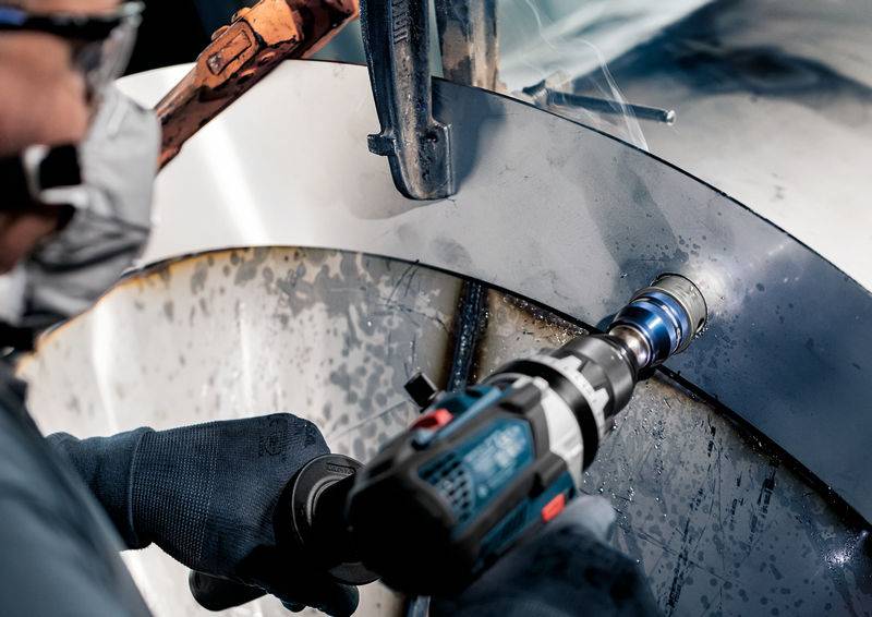 Eine Person arbeitet mit einem Elektrowerkzeug, um ein großes Metallstück zu bohren. Die Umgebung wirkt industriell.