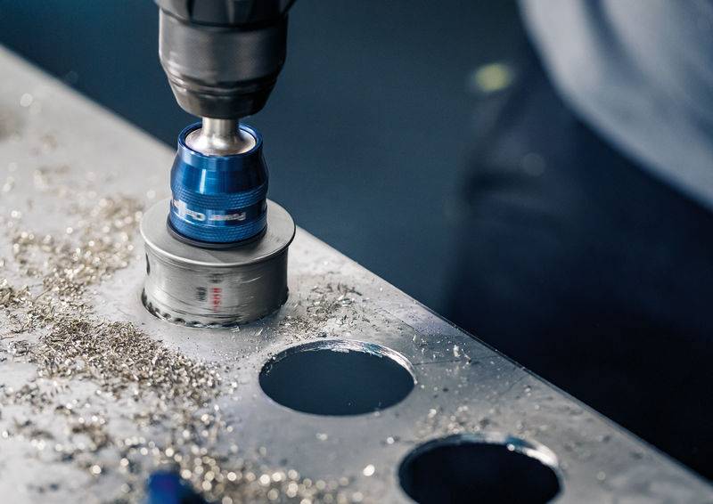 Eine Bohrmaschine schneidet mit einem Lochsägeaufsatz saubere Kreise in ein Metallblech. Metallspäne sind sichtbar verstreut.