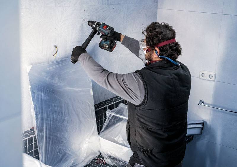 Ein Mann trägt Schutzbrille und montiert mit einer Bohrmaschine ein Zubehörteil an einer gefliesten Badezimmerwand.