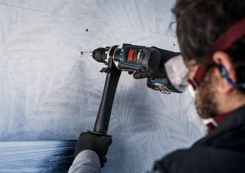 Eine Person bohrt mit einer elektrischen Bohrmaschine ein Loch in die Wand. Sie trägt eine Schutzbrille und eine Maske.