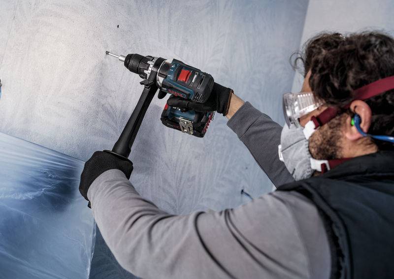 Eine Person trägt Schutzbrille und Staubmaske und bohrt mit einem Akkuschrauber ein Loch in eine Wand.