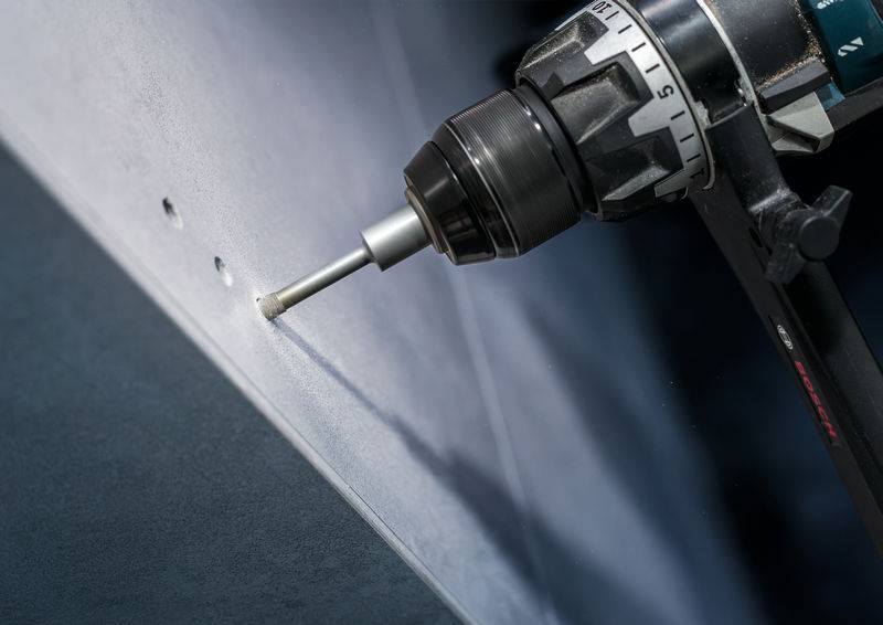Eine Bohrmaschine bohrt Löcher in eine Metallplatte. Nur das Bohrfutter und der Bohrer sind sichtbar, die Löcher haben gleichmäßigen Abstand.