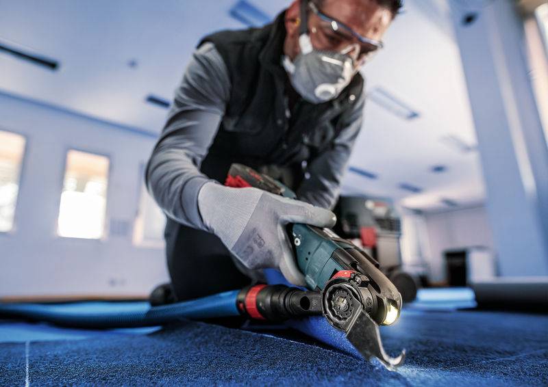 Eine Person schneidet mit einem Werkzeug Schutzmaterial auf dem Boden. Sie trägt Handschuhe, Schutzbrille und Maske in heller Umgebung.